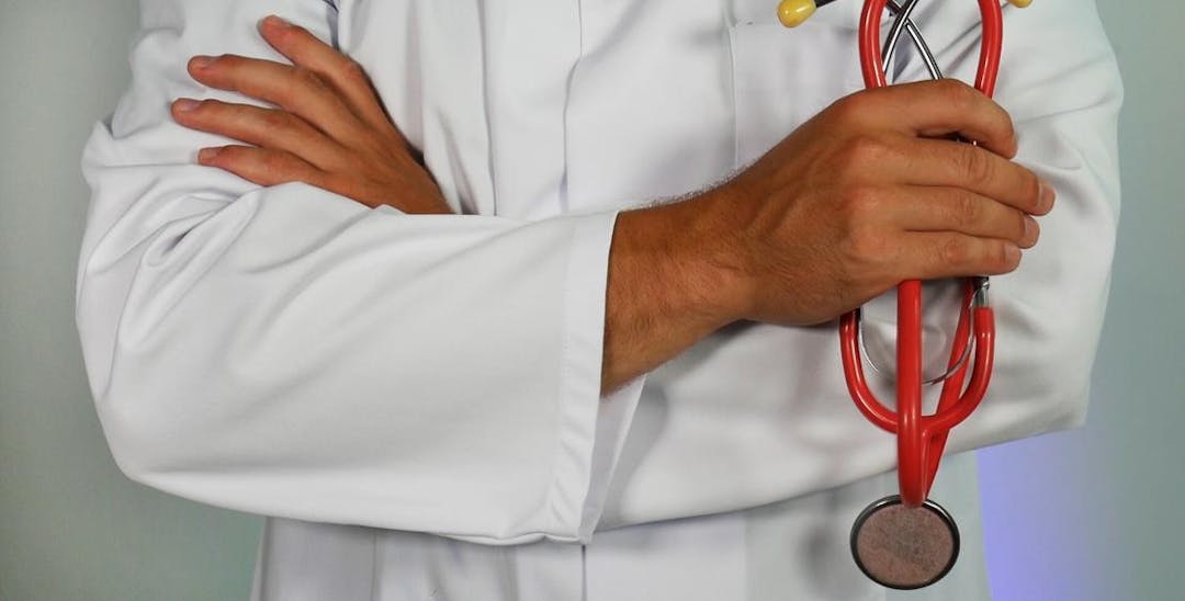 A photo of a torso in a white coat and arms crossed holding a red stethoscope.