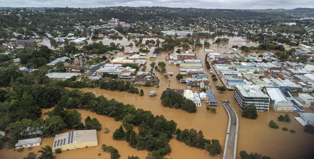 An aerial view of a town during a flood, showing roofs of houses, trees and muddy water covering the streets.