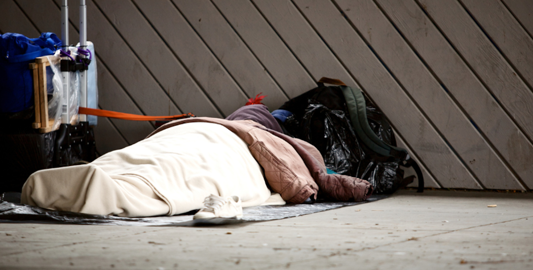 A photo of a person sleeping on the ground outside. They are covered in blankets and surrounded by luggage and other bags.