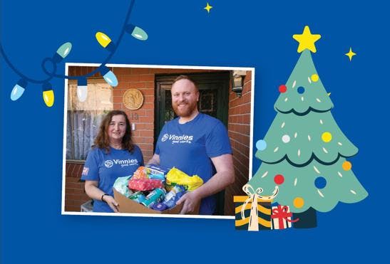 An image with a blue background and a photo of 2 Vinnies WA volunteers holding a box of food donations. Next to the photo is a graphic of a decorated Christmas tree and 2 Christmas presents underneath the tree.