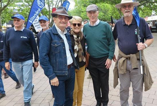 A photo of 4 people standing facing the camera with people walking behind them. They are dressed casually and holding a Vinnies banner.