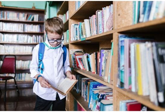 A young boy in a school uniform in a library reading a book. He is wearing glasses and a backpack.
