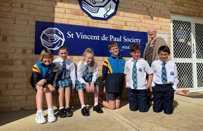 A photo of a group of school children and their teacher standing outside in the sun. Behind them is a wall with a blue sign of St Vincent de Paul Society. The children are wearing school uniforms and they are all smiling at the camera.