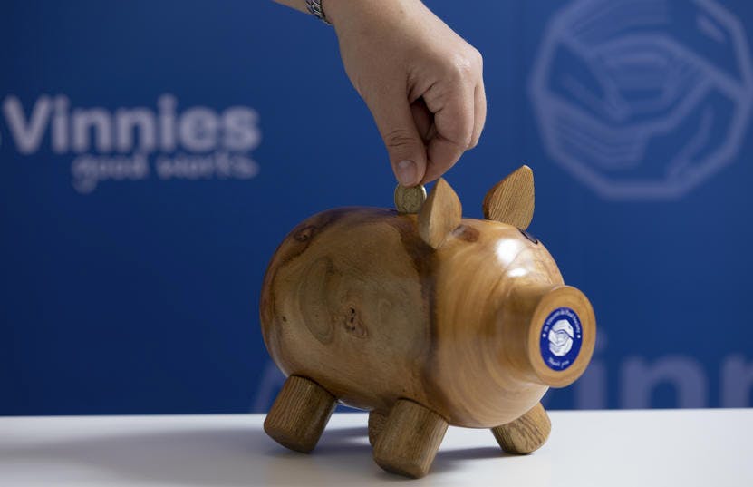 A photo of a hand putting a coin into a wooden piggy bank with a St Vincent de Paul Society sticker on its snout,