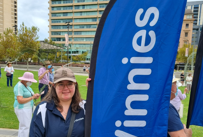 A photo of Claire at an outside event holding a Vinnies banner. She is wearing a Vinnies t-shirt.