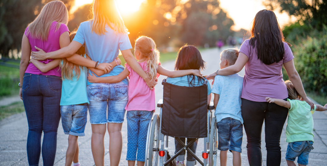 A row of adults and children with their backs to the camera, one in a wheelchair. They have their arms linked and the sun is setting in front of them.