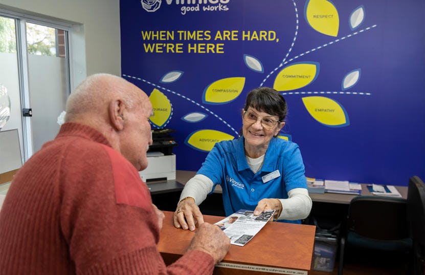 A photo of a Vinnies WA volunteer showing a brochure for an elderly man. They are both sitting down by a desk facing each other and the volunteer is smiling at the man while showing him the brochure. The man has his back against the camera.
