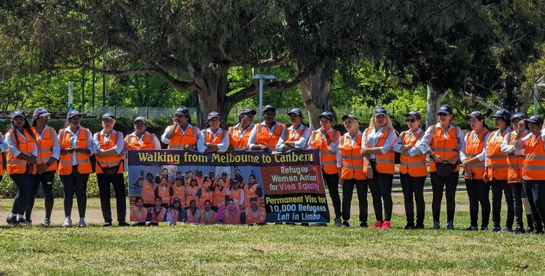A group of women in a line facing the camera, wearing colourful vests and holding a banner saying walking from Melbourne to Canberra.