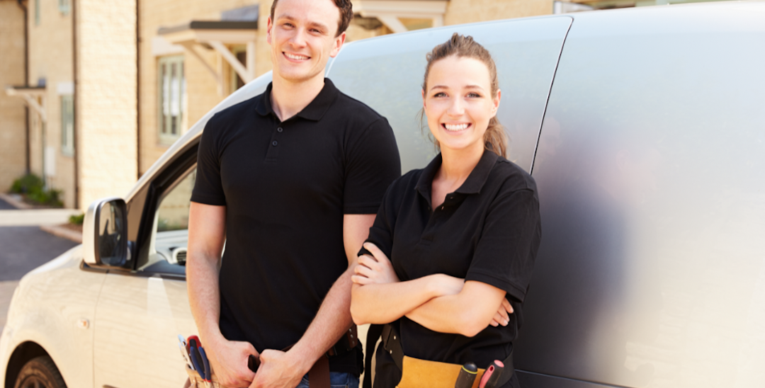 A man and a woman leaning on a white van. They both have tool belts on and are smiling.