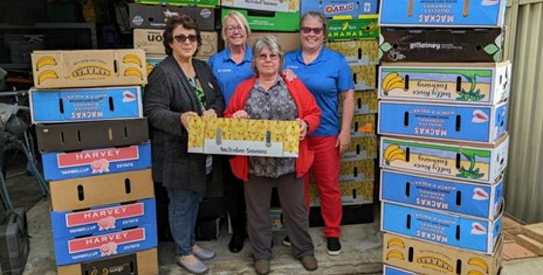 A photo of 4 women holding a box and surrounded by stacks of more boxes in a warehouse. They are facing the camera and smiling.