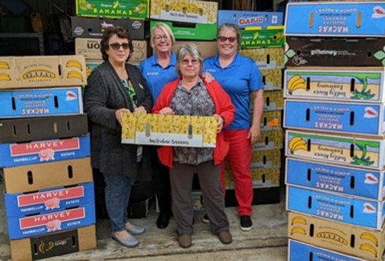 A photo of 4 women holding a box and surrounded by stacks of more boxes in a warehouse. They are facing the camera and smiling.