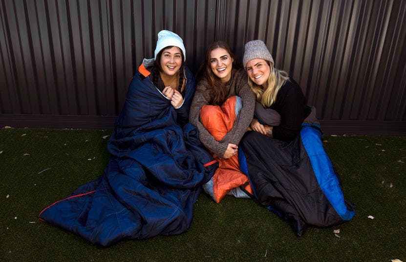 A photo of 3 participants of the Vinnies WA Tough Night Out sitting on the ground in a sleeping bag each. 2 of them are wearing hats and they all have warm clothes on. They are smiling at the camera.