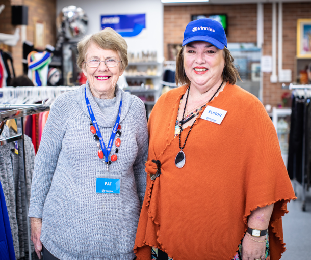 Two smiling women who volunteer for Vinnies shops are standing in a Vinnies shop.