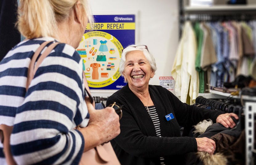 A photo of a Vinnies WA volunteer working at a Vinnies shop. The volunteer is smiling at a customer who has her back to the camera, while the volunteer has her hands on a rack of clothes. 