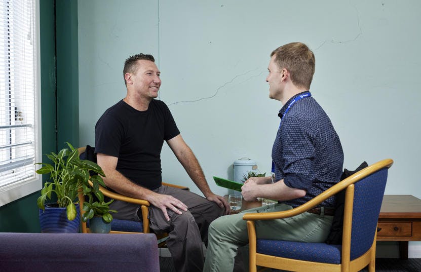 A photo of a man and a Vinnies WA volunteer sitting down in chair opposite each other having a conversation. The volunteer is holding a green brochure while the man is smiling at him. 
