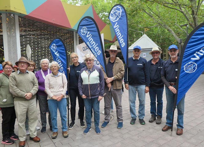 A photo of a group of men and women standing, in casual clothes and some in hats, holding Vinnies banners and posing for the camera.