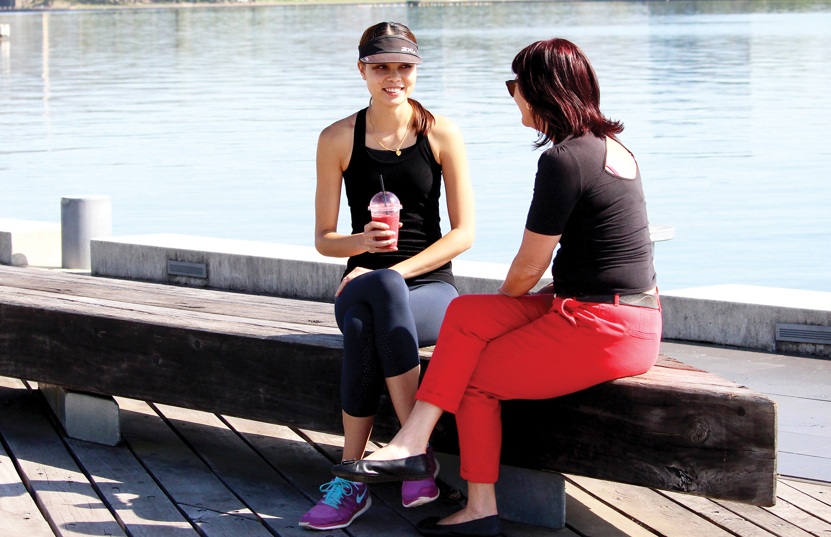 Two women sitting by a lake drinking iced coffees and chatting.