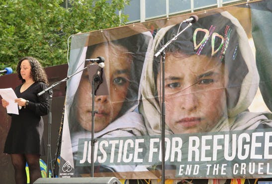 A phot of a woman in a black dress with long black, curly hair talking into a microphone, on a stage, at an outdoors rally. The backdrop features a picture of 2 girls and reads Justice for Refugees.