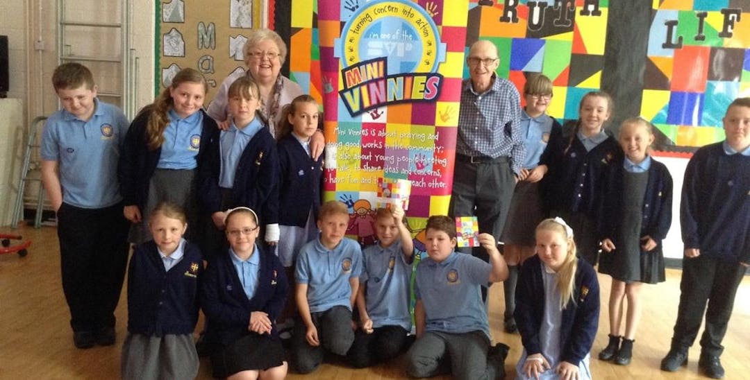 A group of children in school uniform and two adults standing and sitting in front of a Mini Vinnies banner in a school hall and smiling at the camera.
