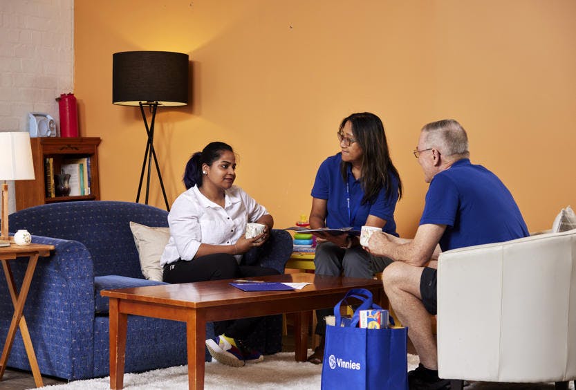 A photo of 2 Vinnies WA volunteers sitting down with a woman offering her assistance. The volunteers are wearing blue t-shirts and wearing lanyards.