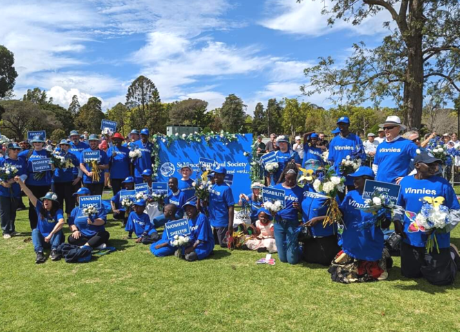 A photo of Vinnies volunteers in QLD in a park, in blue t-shirts with signs and banners on green grass in a park.