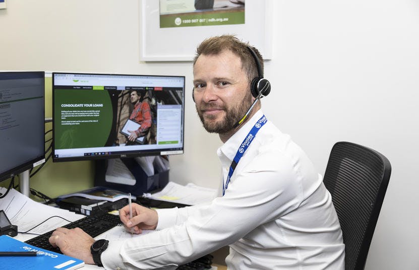 A photo of a Vinnies WA volunteer at the callcentre. He is sitting by a desk in front of 2 computer screens and wearing headphones. He is holding a pen and facing the camera.