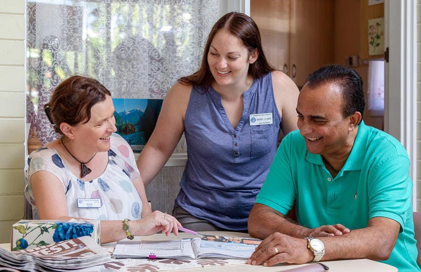 A photo of 2 volunteers assisting a man in need of Vinnies WA's services. One volunteer and the man are sitting down by a table, while the second volunteer is standing behind them leaning against their chairs. They are all smiling, looking at each other or the information brochure on the table.