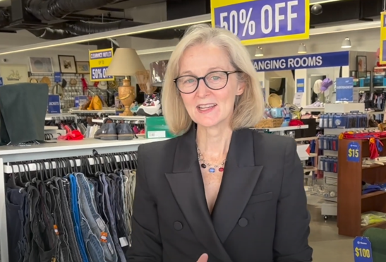 A photo of the Chief Executive Director Susan Rooney at a Vinnies WA shop. She is centre of the photo with clothes racks and clearances signs in the background. She is facing the camera while talking.