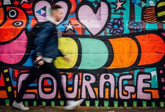 A photo of a man walking fast past a wall covered with colourful graffiti artwork and the word "COURAGE".
