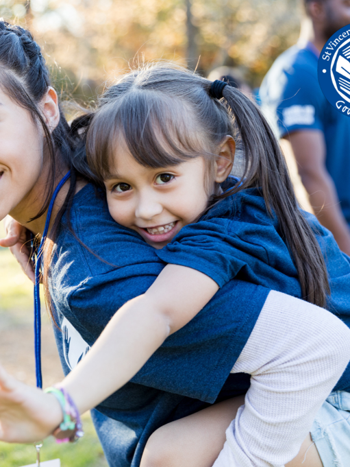 A women wearing a Vinnies blue shirt is carrying a small children aged approximately six years old on her back.  Both are smiling. There are two men in the distance also in Vinnies blue shirts. 
