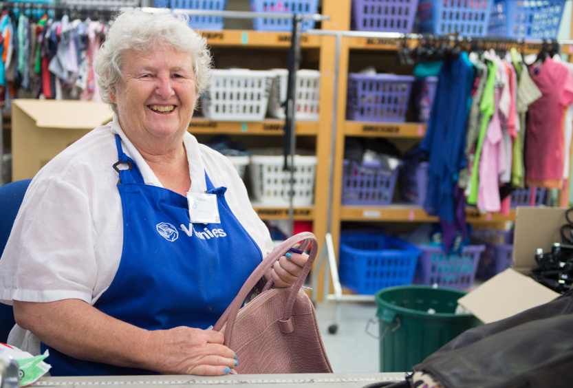A photo of a lady in a Vinnies shop wearing a blue Vinnies apron and name badge, holding a handbag. In the background are rows of shelves with baskets on them and clothes on racks. She is facing the camera and smiling.