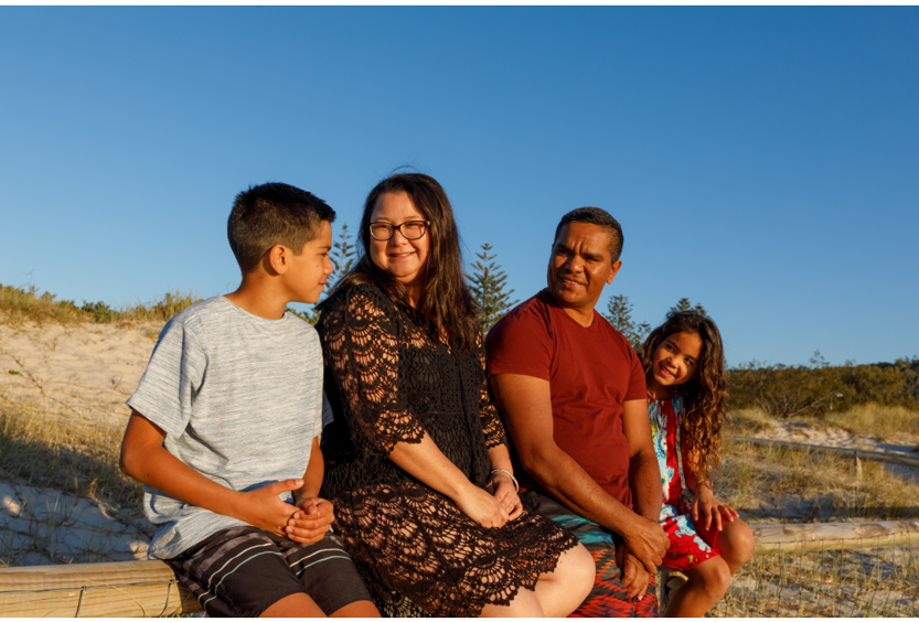 A family of 4 sitting on a log near sand dunes, they are all looking at the boy and smiling. The sky behind them is very blue.