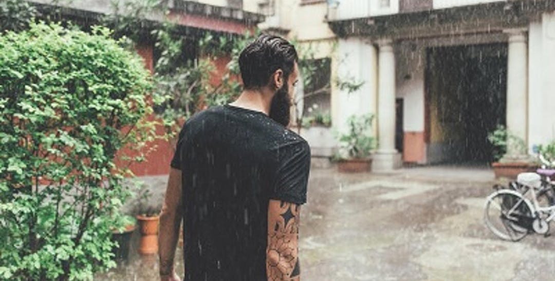 A photo of a man with a beard and tattoos walking away from us towards a door. It is raining.
