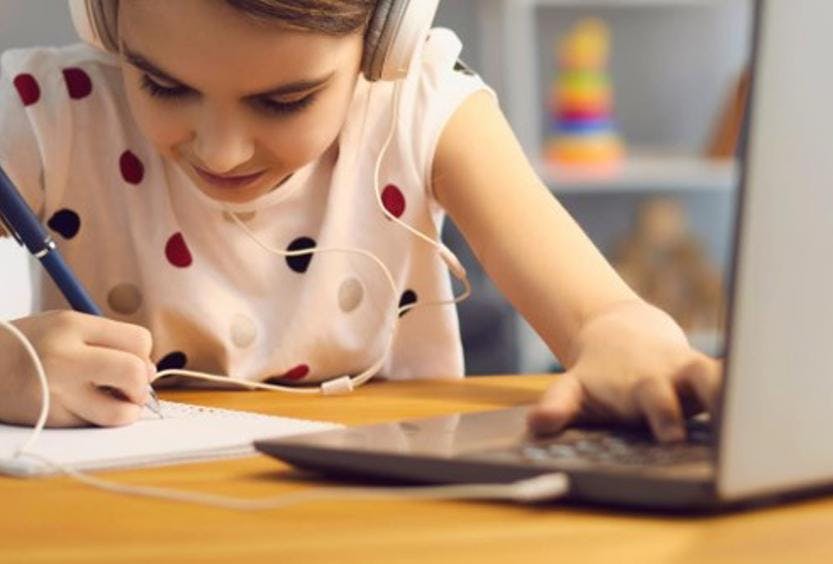 A picture of a young girl in a spotty t-shirt, wearing headphones and writing on a pad of paper. She has a laptop in front of her.