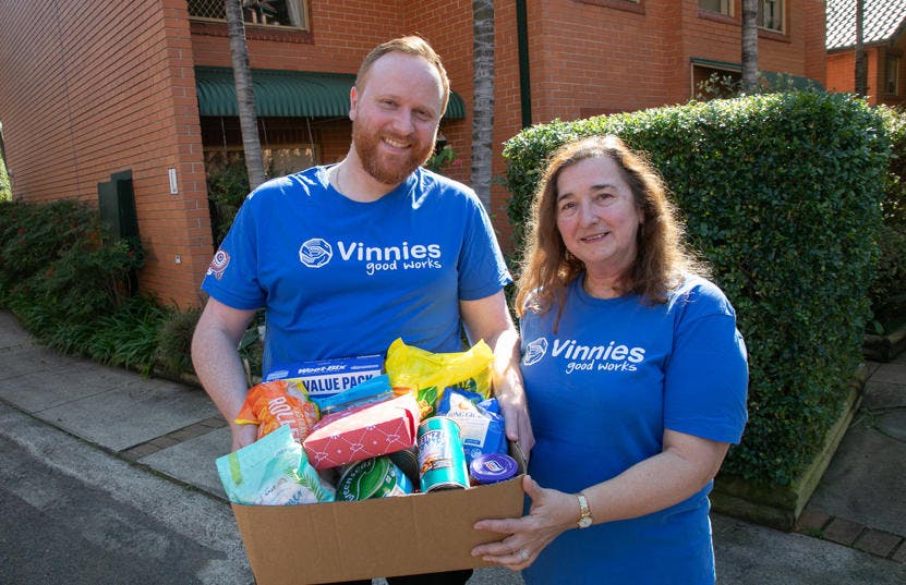 2 people wearing blue Vinnies t-shirts, holding a box of donated food and smiling at the camera.