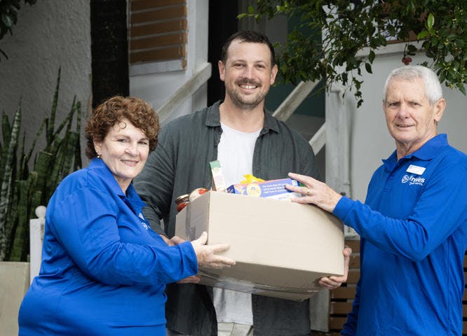 2 men and a woman holding a box of food. One man and the woman are wearing Vinnies blue tshirts. They are all smiling and facing the camera.