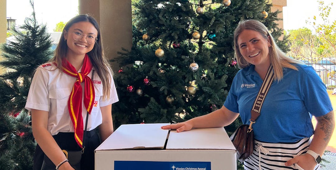 A photo of 2 women standing in front of a decorated Christmas tree with a large, white box between them for donated items. One is a Vinnies WA staff wearing a blue Vinnies pique jersey and the other is a volunteer in a working uniform. They are both smiling at the camera.