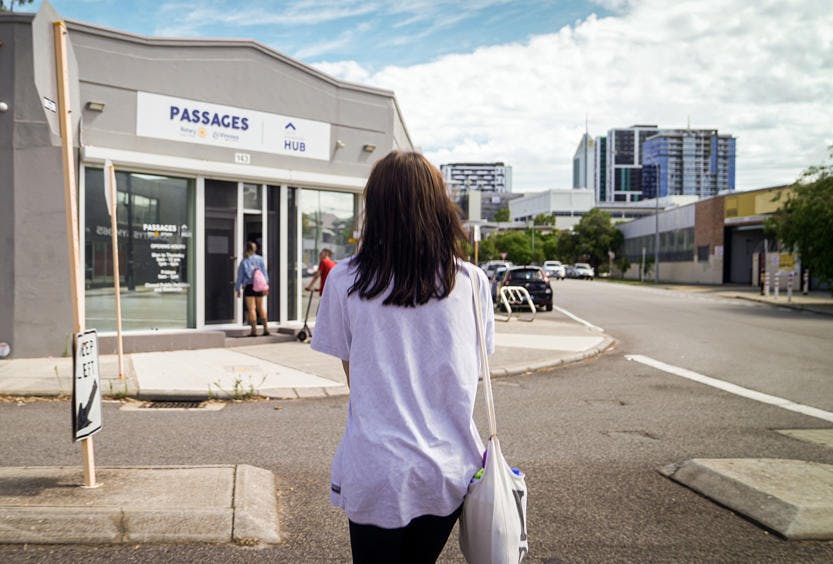 A photo of a woman's back as she is crossing the road walking towards a Passages Youth Engagement Hub. She is carrying a bag and wearing a purple T-shirt.