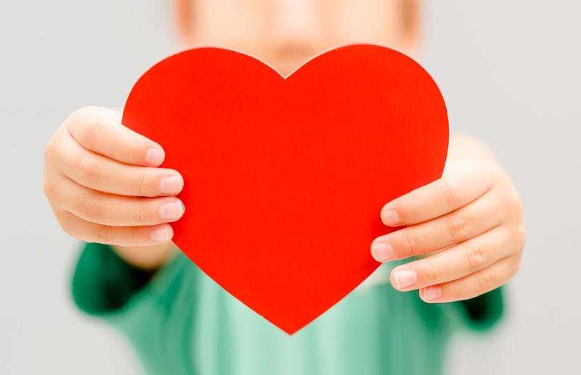 A photo of a child holding up a red heart image in front of the camera. The child is wearing a green t-shirt which is not in focus, while the hands and the heart image is in focus.