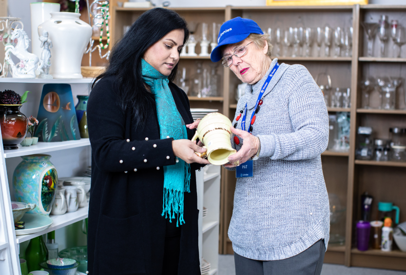 A photo of 2 women in a Vinnies shop looking at the bottom of a jug. They are surrounded by shelves of china and glass and 1 woman is wearing a Vinnies cap.