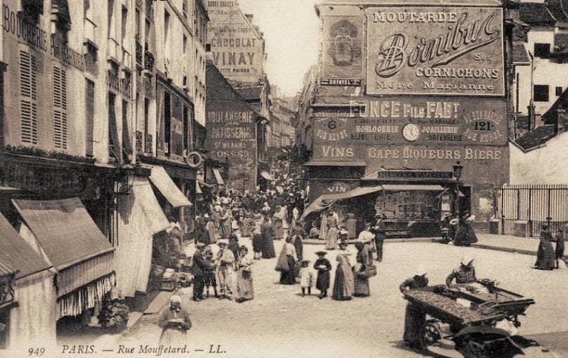 A sepia-washed photo shows a street in Paris in the 1980s. It includes two- and three-story buildings and people walking in the street.
