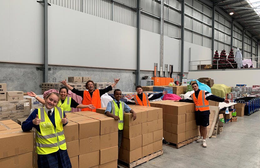 A photo of a group of Vinnies WA Volunteers at the Vinnies WA distribution centre. They are standing among stacked, closed boxes for donation and are all wearing yellow or orange vests. They are all smiling and the camera.