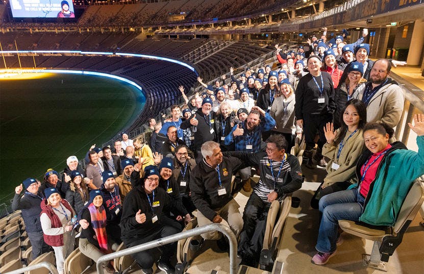 A photo of the crowd of participants from the CEO Sleepout at Optus Stadium. They are either standing up or sitting down, while facing the camera and smiling.
