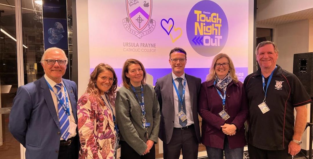 A photo taken of a group of 6 adults, 3 men and 3 women that are all smiling at the camera. In the background is a projector screen showcasing an image of the Tough Night Out logo and the logo of Ursula Freyne Catholic College. Everyone is wearing Vinnies WA lanyards.