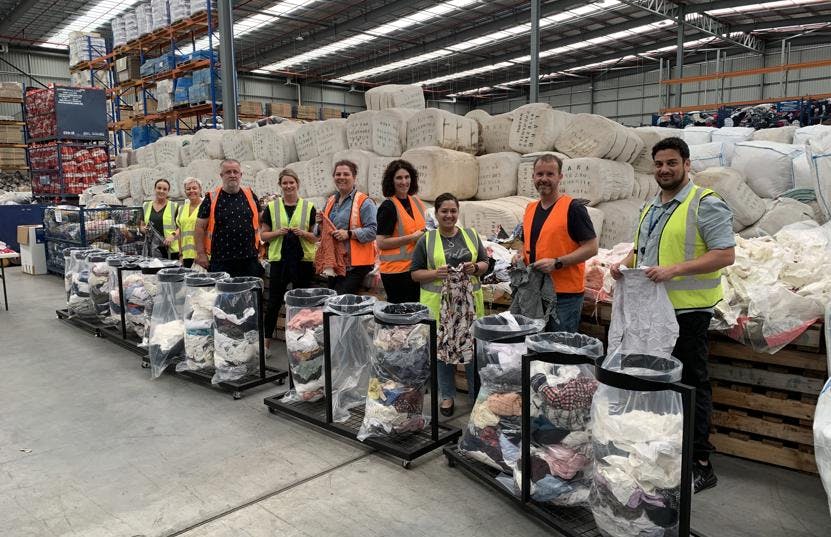 A photo of 9 Vinnies WA volunteers sorting clothes donations at a storehouse. They are all wearing yellow or orange vests and have on bag each filled with clothes.