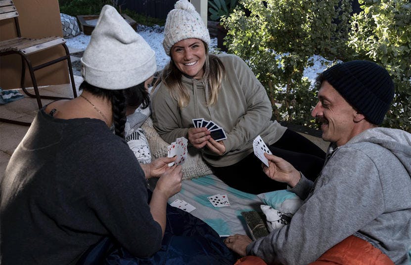 A photo of 3 participants of the Vinnies WA Tough Night Out sitting outside playing a game of cards. They are all wearing hats and warm jumpers. One woman is facing the camera smiling, while the other two has their backs towards the camera. 