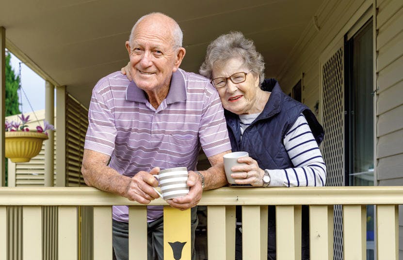 A photo of an elderly couple standing on their porch leaning on the fence and drinking coffee. They are both looking into the camera and smiling. The woman has her arm around the man and leans her head against his shoulder.