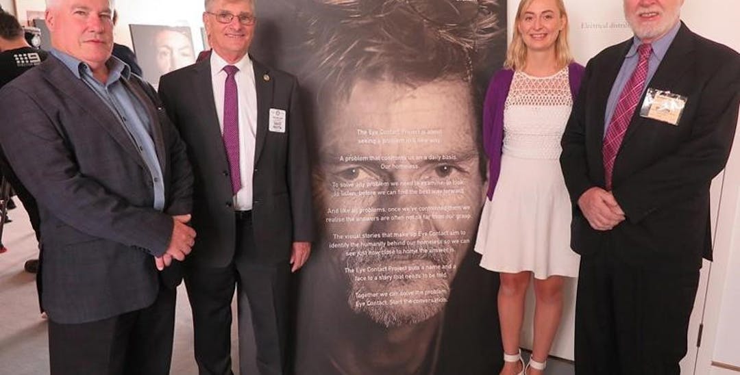 A photo of 4 people standing in a room, in front of a banner with a photo of a man on it. They are all looking at the camera.