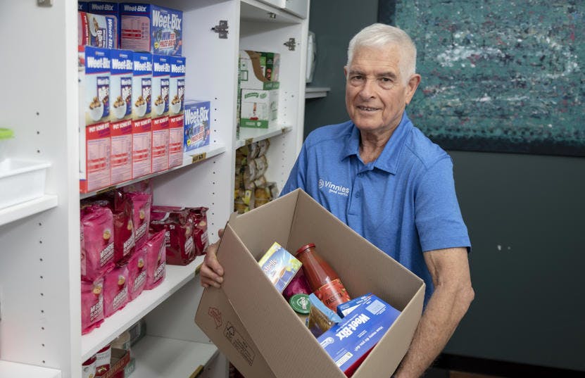 An elder Vinnies WA volunteer holding a box of food while standing next to a grocery shelf. The man is wearing a blue pique jersey and smiling at the camera. 