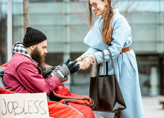 A photo of a woman in smart work clothes handing food to a man sitting on the street in a sleeping bag. wearing a beanie. His cardboard sign says jobless.